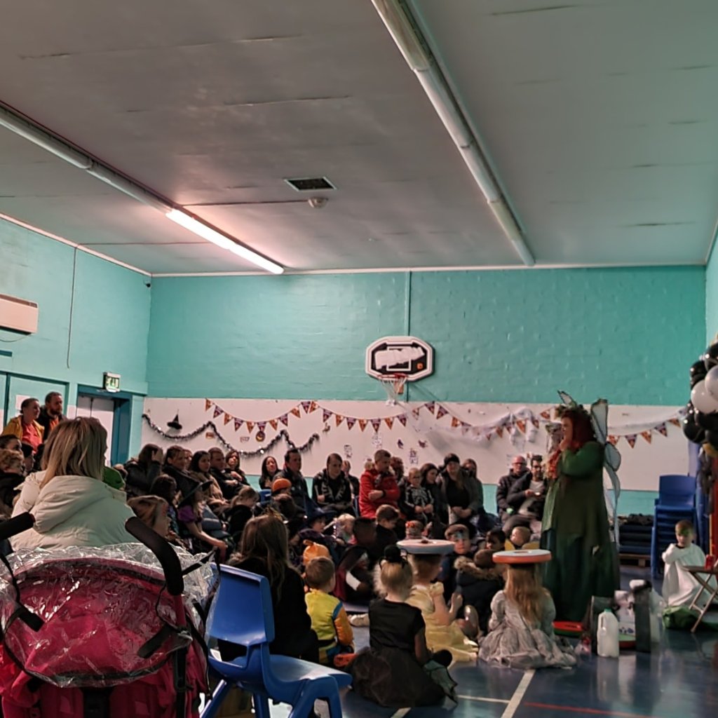 A photograph of the school hall at Middleton Park Primary - Pauline stands on the right of the photo dressed as a fairy in green robes and with blue fairy wings.  She is facing lots of children and their grown ups who are listening to the story