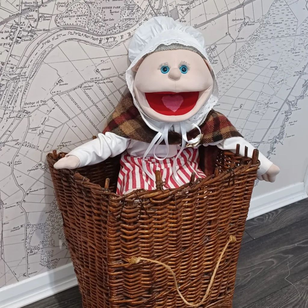 A photo of an elderly female puppet dressed as a traditional fishwife from the north east of Scotland.  She is wearing a white mutch cap on her grey hair, a cloak made of checked woollen material a white blouse and a red and white striped apron.  She is sitting inside an antique woven creel which is just the perfect size to hold her. 