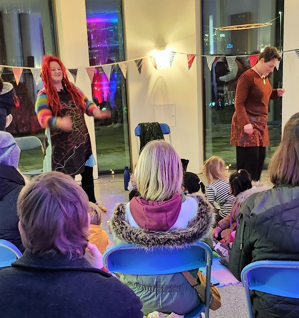 Pauline Cordiner and Lindsey Gibb tell stories at Spectra Light Festival 2023. Pauline gestures at the crowd while Lindsey laughs. 