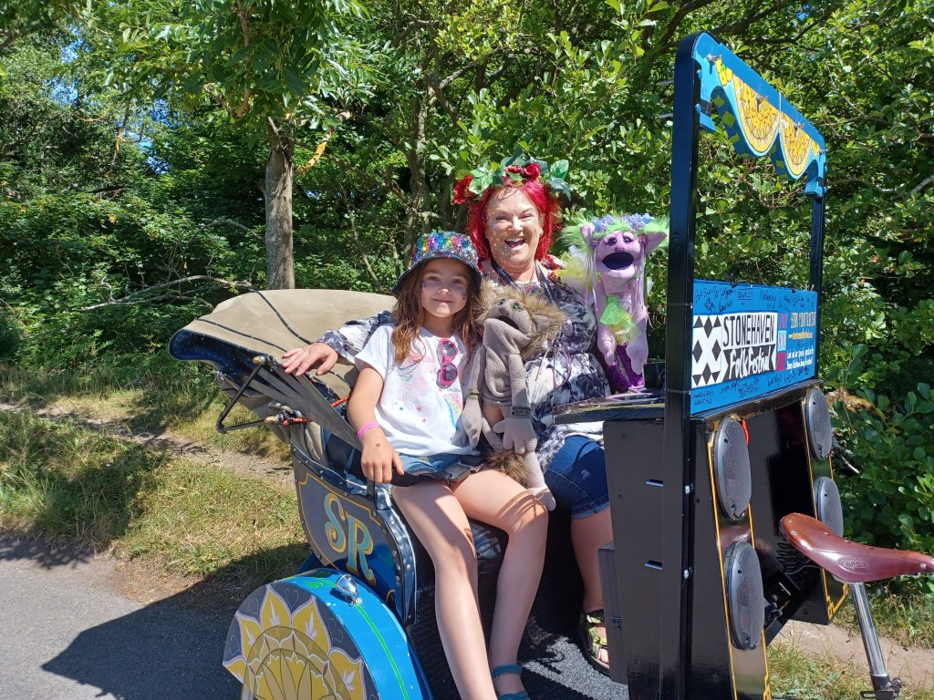 Pauline, her daughter, and Fuzzy and Fizzy - two fairy puppets - sit in a Rickshaw in the sunshine smiling at the camera
