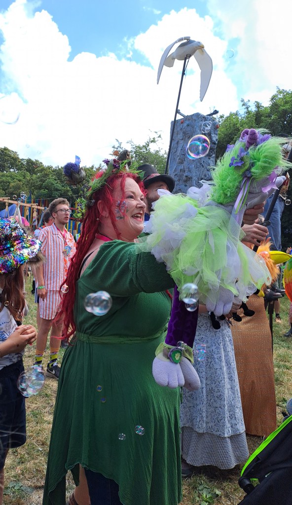 A photo of Pauline surrounded by colourful entertainers at the opening of the Kidz Field at this year's Glastonbury Festival.  She is holding Fairy Puppet, Fizzy the Fairy who is greeting everyone coming in. 