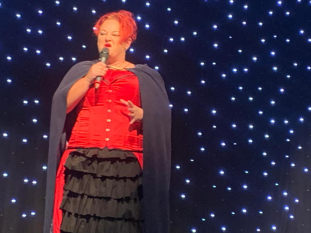 Pauline telling stories on stage at the Beach Ballroom. Wearing black and red dress outfit with cloak.  She looks at the audience telling the story with a microphone in hand. 
