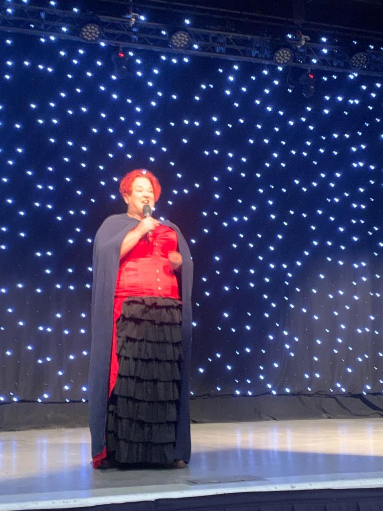 Pauline telling stories on stage at the Beach Ballroom. Wearing black and red dress outfit with cloak.  She looks at the audience telling the story with a microphone in hand. 