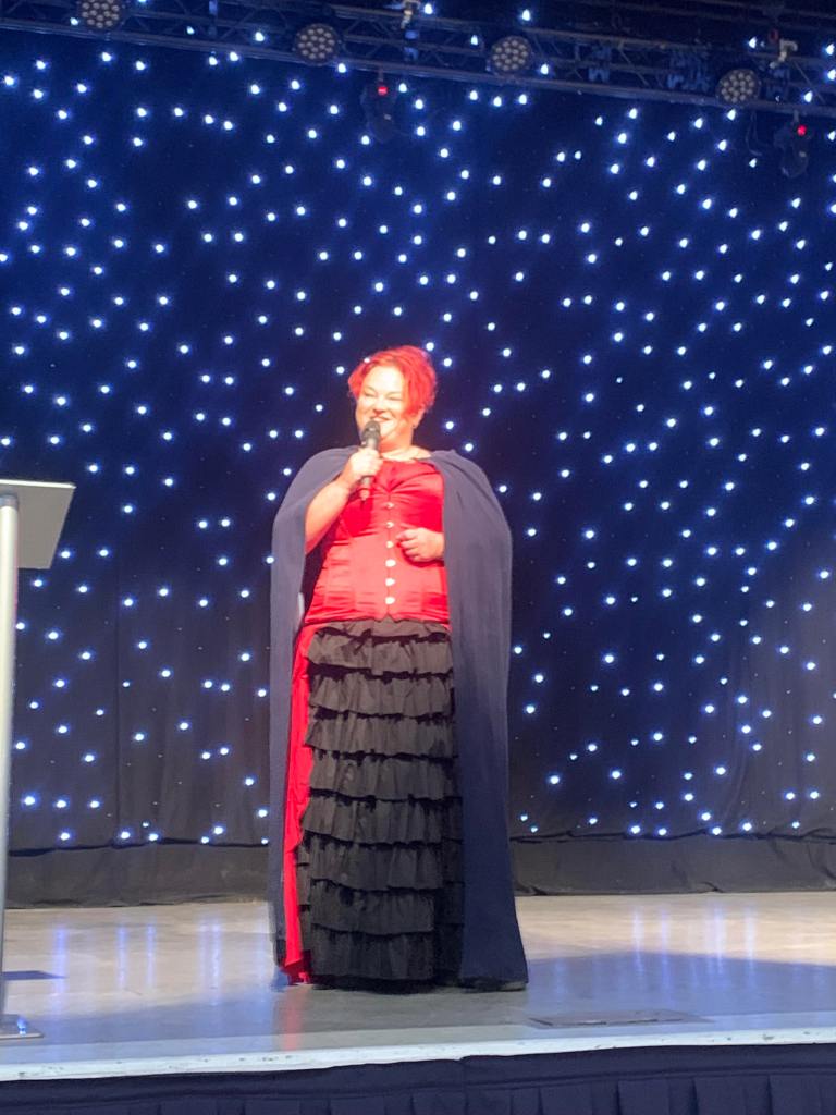 Pauline telling stories on stage at the Beach Ballroom. Wearing black and red dress outfit with cloak.  She looks at the audience telling the story with a microphone in hand. 